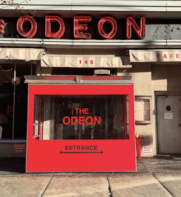 Bright red entrance booth with THE ODEON and ENTRANCE written on it stands outside a building, near Sylvias, with a large neon ODEON sign above and the address number 145 visible. Sunlight casts shadows on the sidewalk.